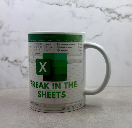 A white ceramic mug featuring a Microsoft Excel spreadsheet theme with green and gray details and the text 'FREAK IN THE SHEETS' on a light gray marble surface.
