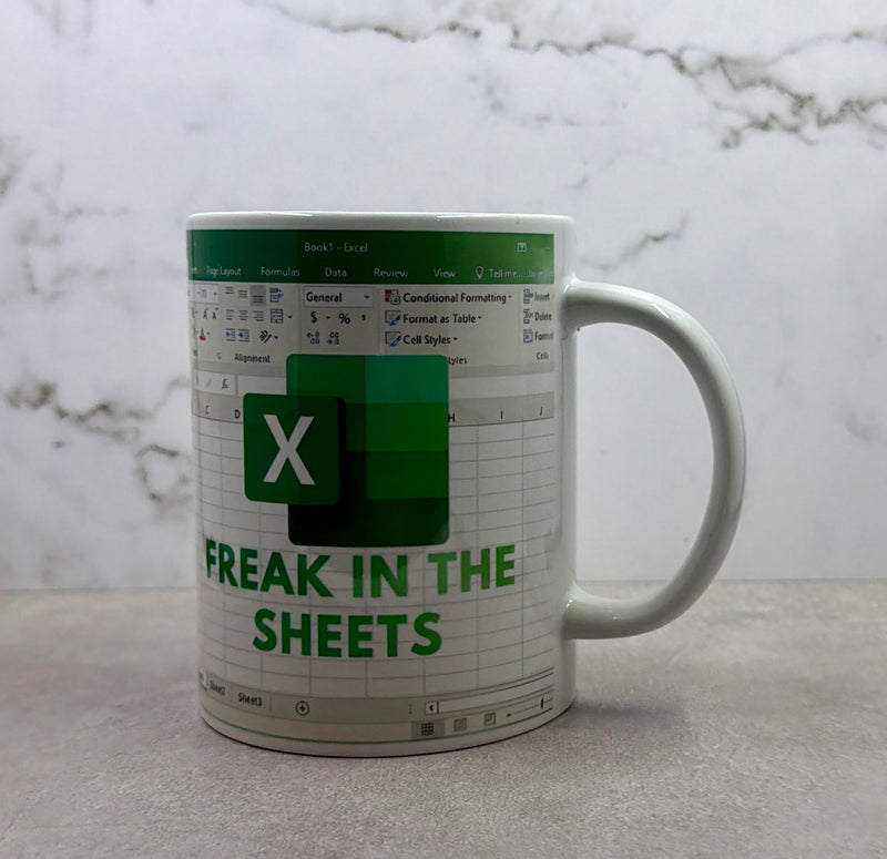 A white ceramic mug featuring a Microsoft Excel spreadsheet theme with green and gray details and the text 'FREAK IN THE SHEETS' on a light gray marble surface.