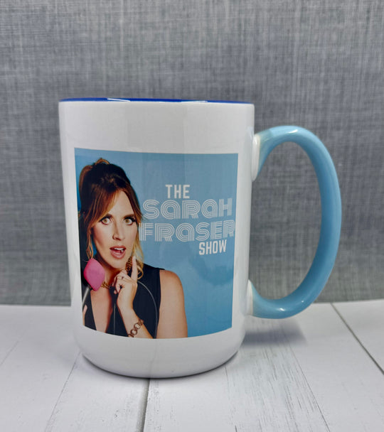 A white ceramic mug with blue beveled edge and handle, featuring an image of a woman and the text 'THE SARAH FROSTER SHOW'. There are pink accents on the mug.